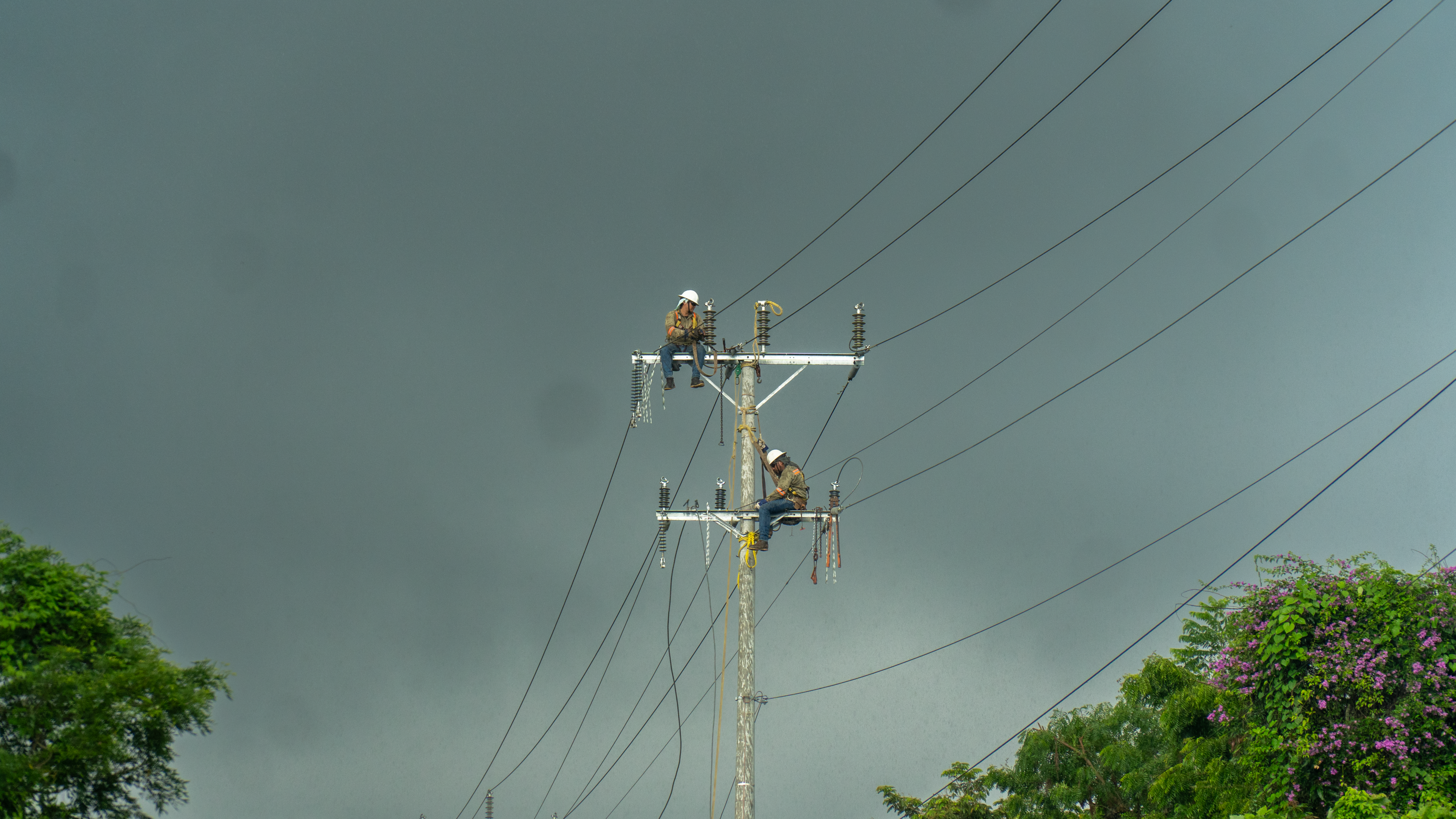 Dos trabajadores de servicios públicos con cascos y arneses de seguridad están sentados en una línea de servicio público o poste de electricidad. Están reparando cables eléctricos de alto voltaje. En el fondo, un cielo oscuro y tormentoso.
