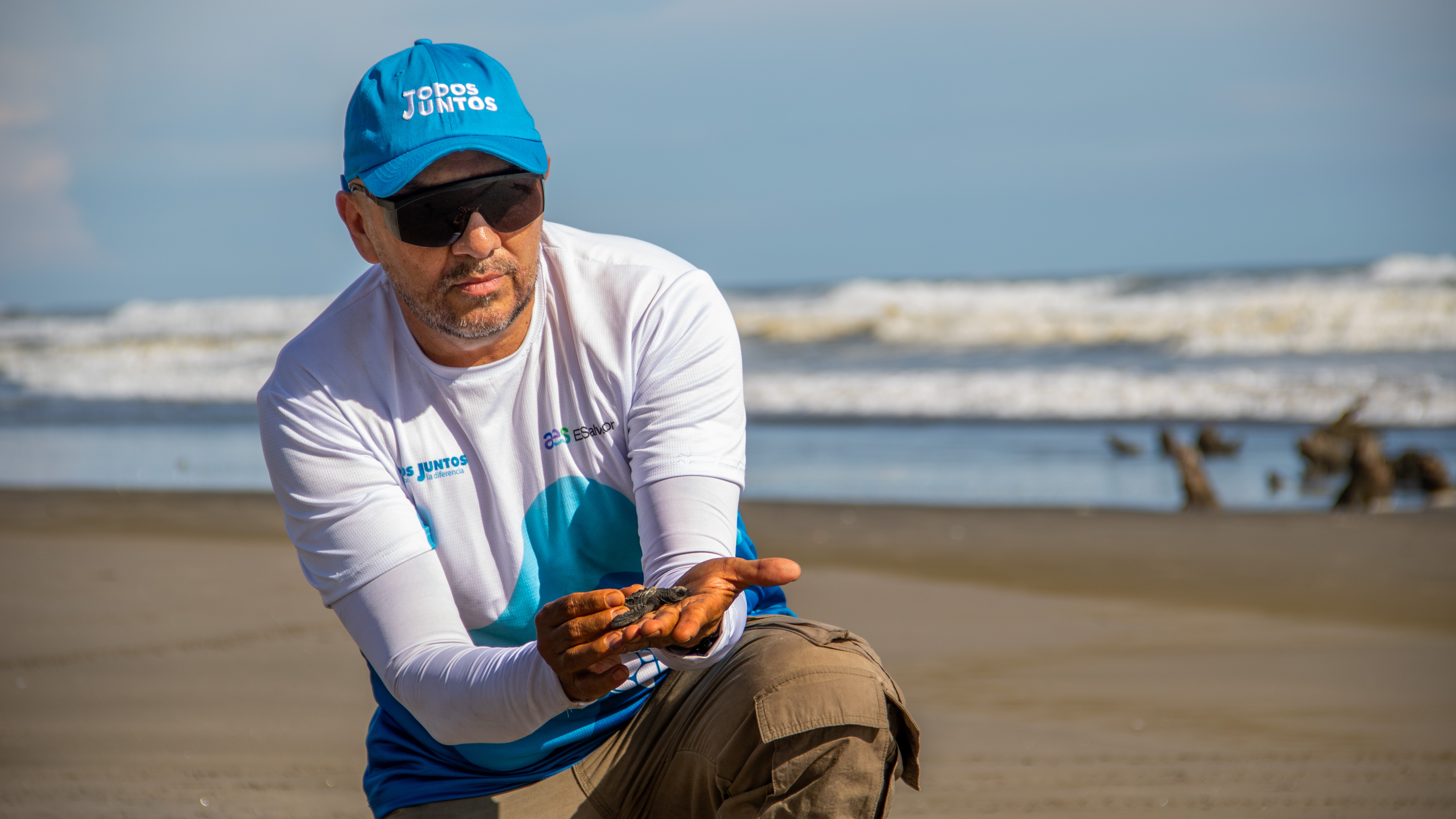 Un hombre, que usa una gorra azul con la frase "Todos Juntos", una camiseta blanca con detalles azules y gafas de sol oscuras, está agachado en una playa arenosa. Sostiene cuidadosamente una pequeña tortuga marina bebé en sus manos. En el fondo, el océano con olas rompiendo es visible bajo un cielo ligeramente nublado.