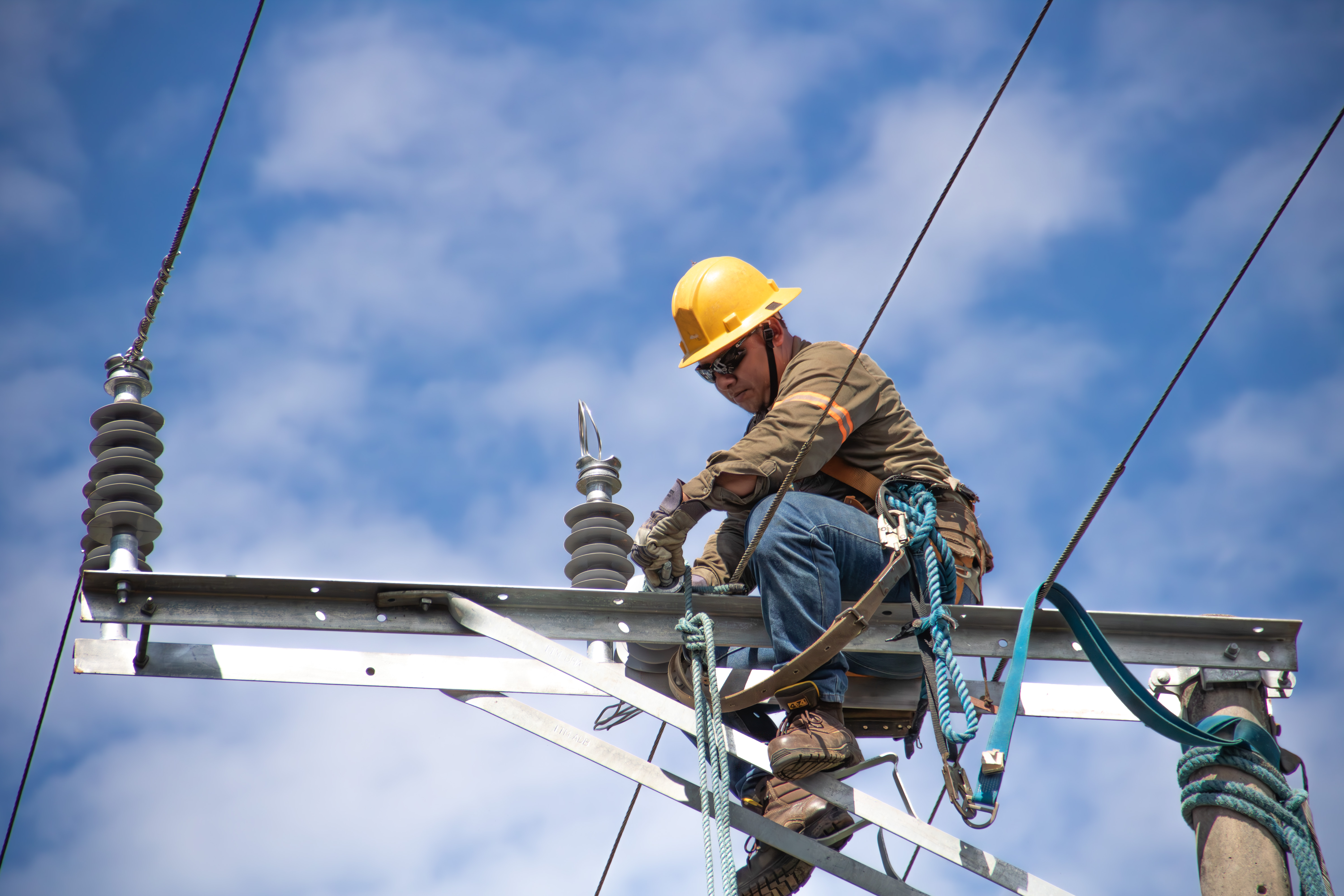 Un ingeniero eléctrico con casco y equipo de seguridad trabaja en la parte superior de un poste de electricidad, realizando mantenimiento en las líneas de alto voltaje. Se le ve enfocado en sus tareas bajo un cielo azul con nubes dispersas, asegurando la infraestructura eléctrica.