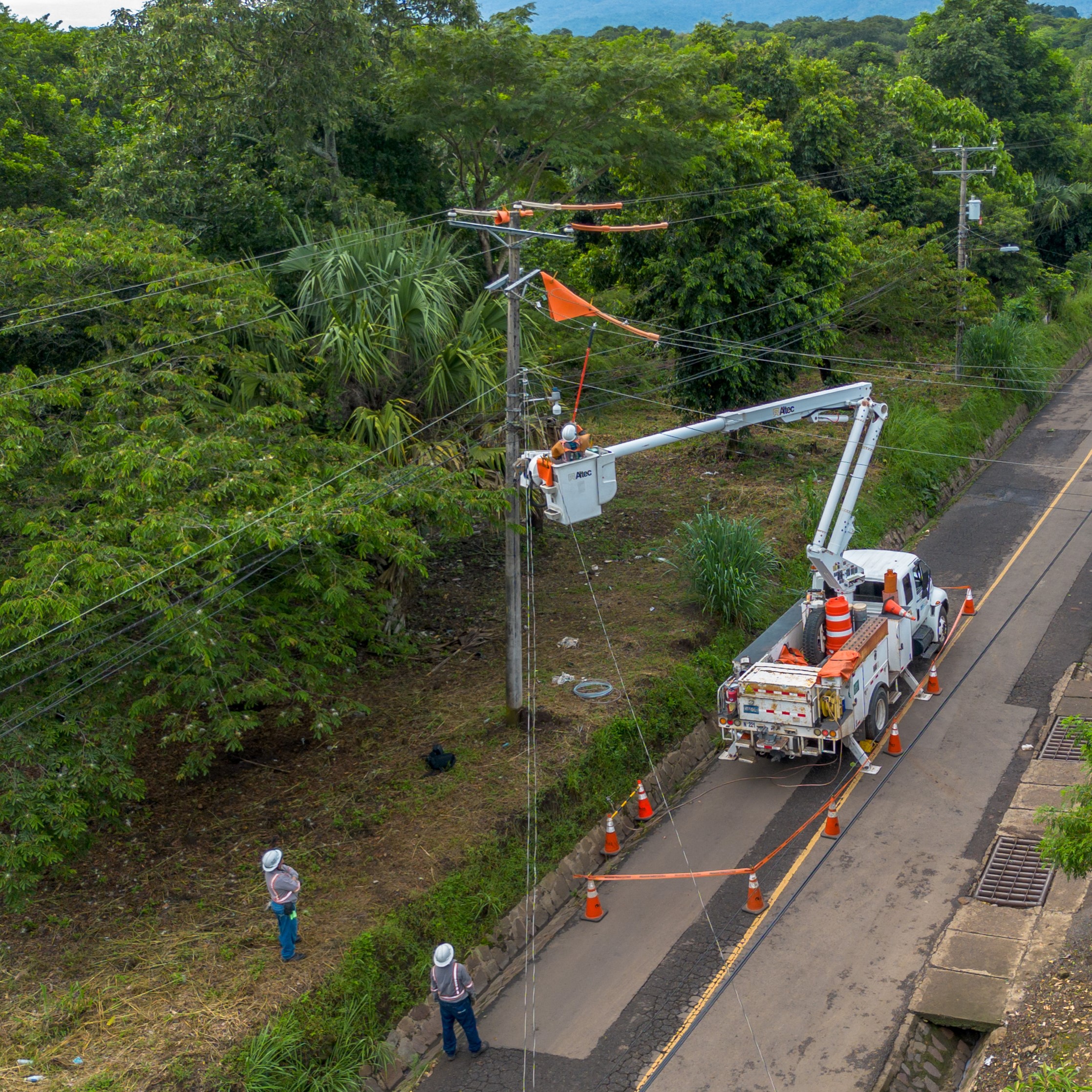 Vista aérea de un equipo de trabajadores eléctricos realizando mantenimiento en un poste de energía. Un trabajador está elevado en la canasta de una grúa, operando cerca de los cables, mientras dos colegas lo observan desde el suelo. El entorno es una carretera rural bordeada por densa vegetación y postes de electricidad, destacando la infraestructura energética en un paisaje natural. 