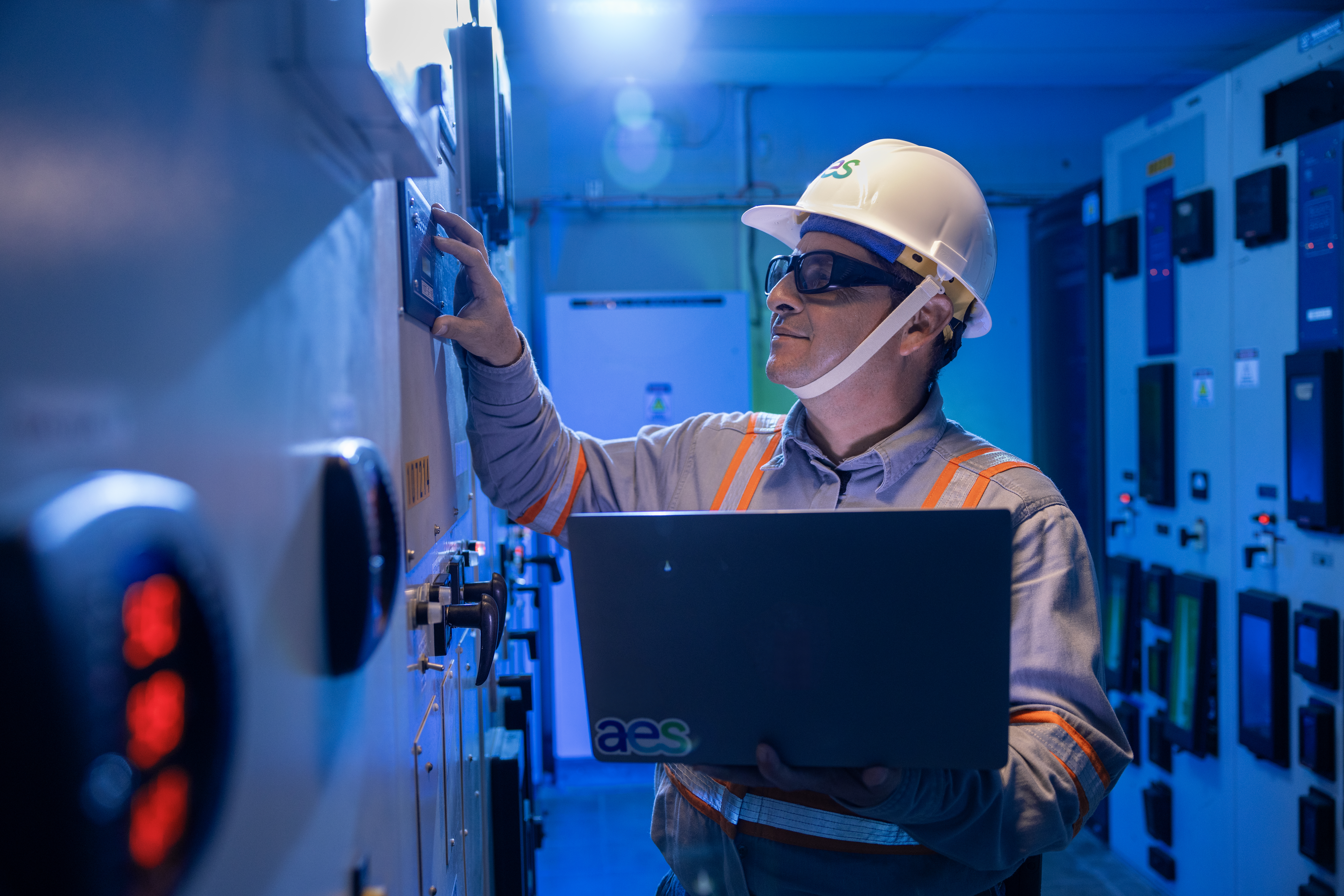 Técnico de AES con casco blanco y lentes de seguridad trabaja en una sala de control iluminada con luz azul. Sostiene una computadora portátil con una mano mientras ajusta controles en un panel eléctrico industrial. El ambiente sugiere un entorno de alta tecnología y monitoreo energético.