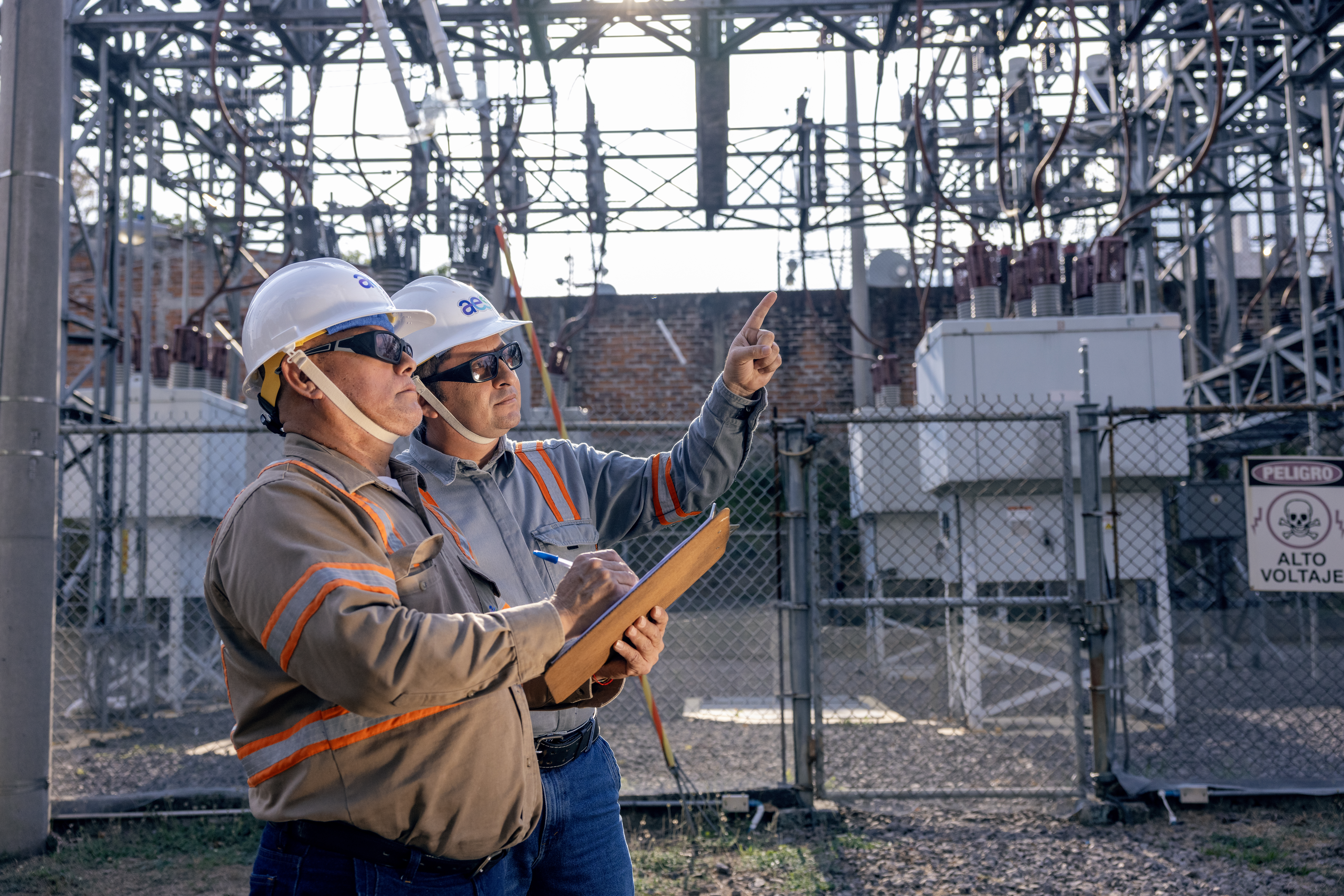 Dos técnicos de AES realizan una inspección en una subestación eléctrica al aire libre. Uno de ellos señala hacia la estructura metálica de alta tensión mientras el otro toma notas en una tablilla de madera. Ambos visten uniformes de seguridad, cascos blancos y lentes oscuros bajo la luz del día.