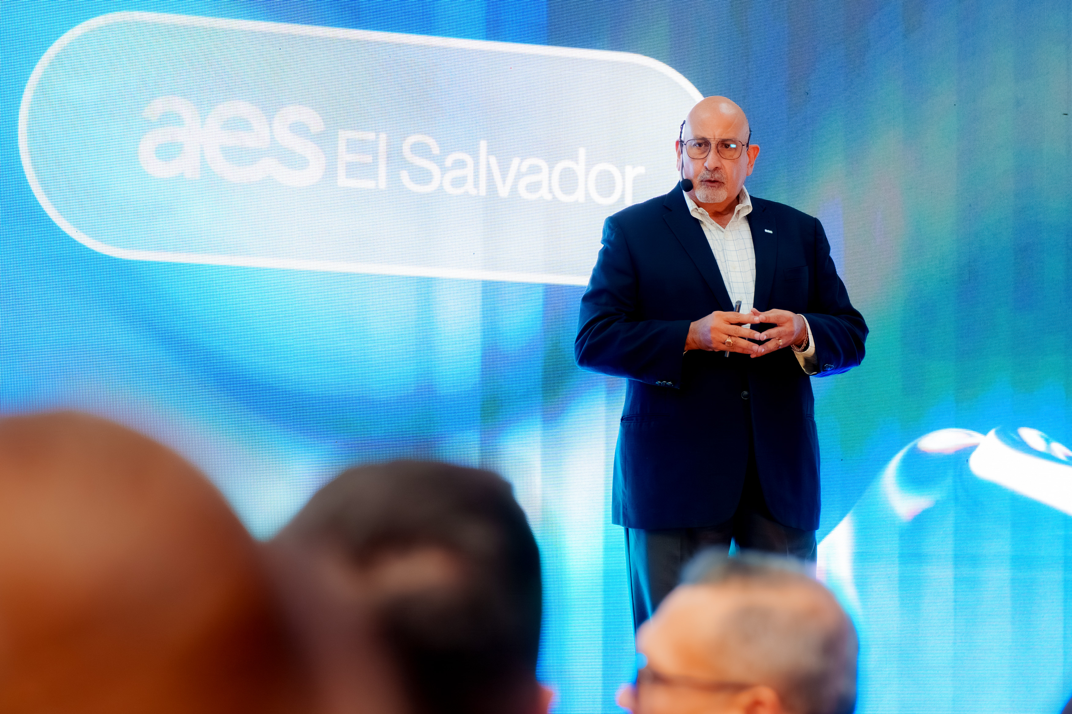 A man in a suit speaks on stage with a microphone headset in front of a large screen displaying the AES El Salvador logo, addressing an audience.