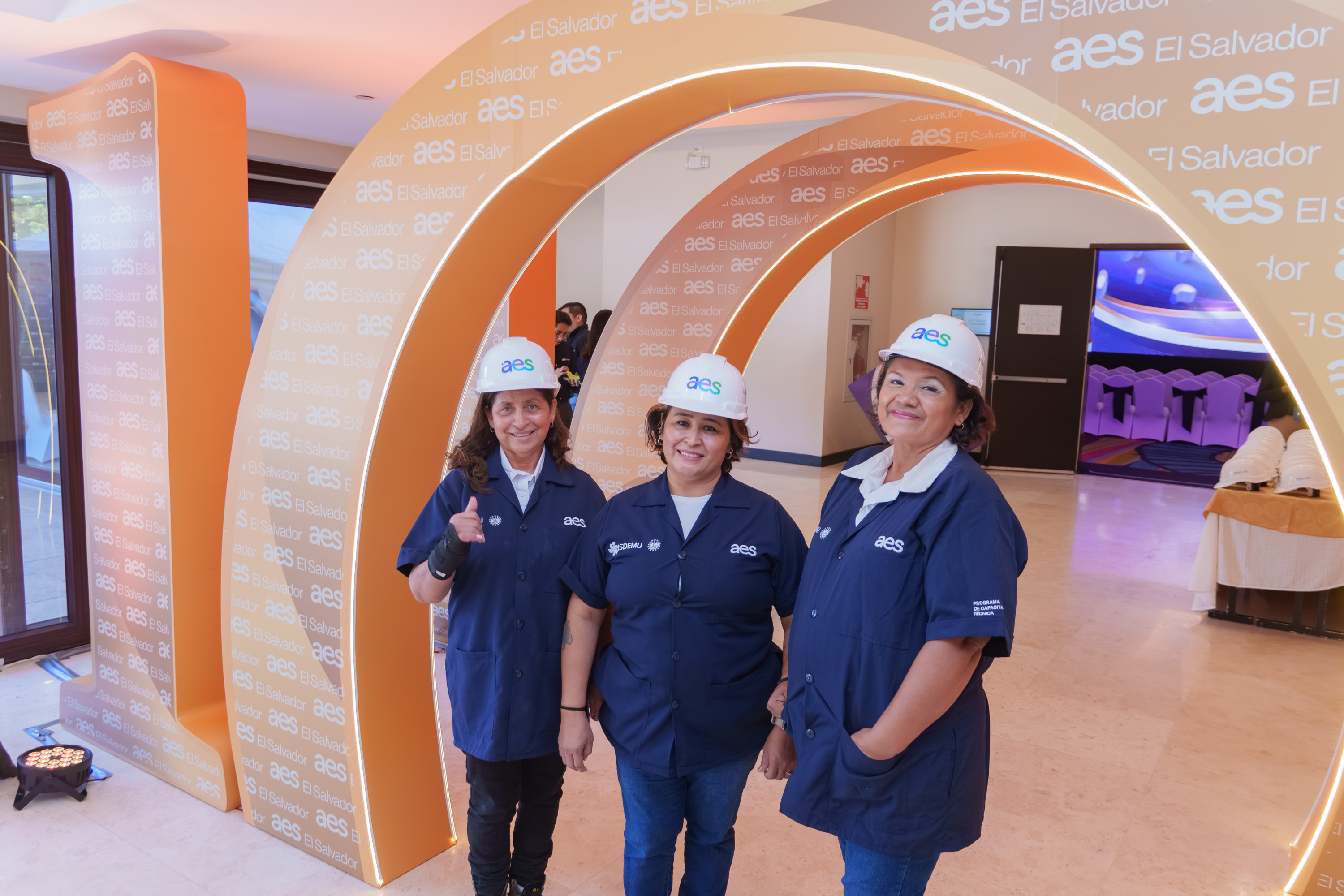 Three women wearing AES hard hats and uniforms stand smiling under a large orange arch with 'AES El Salvador' text at an indoor event.