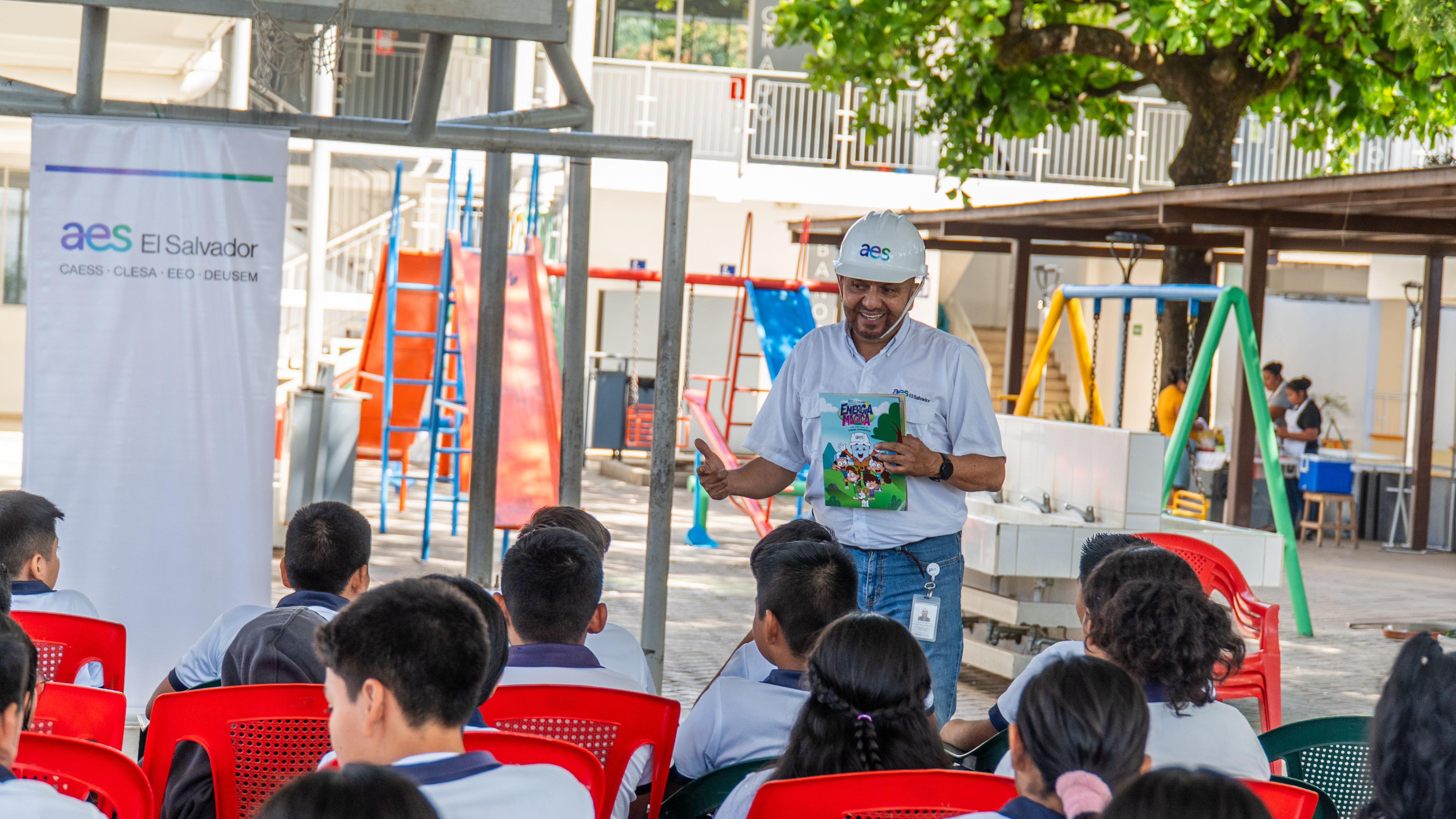 A man wearing a white hard hat and holding an educational booklet speaks to a group of students seated outdoors. The students are sitting on red chairs, and playground equipment and a banner reading 'aes El Salvador' are visible in the background.