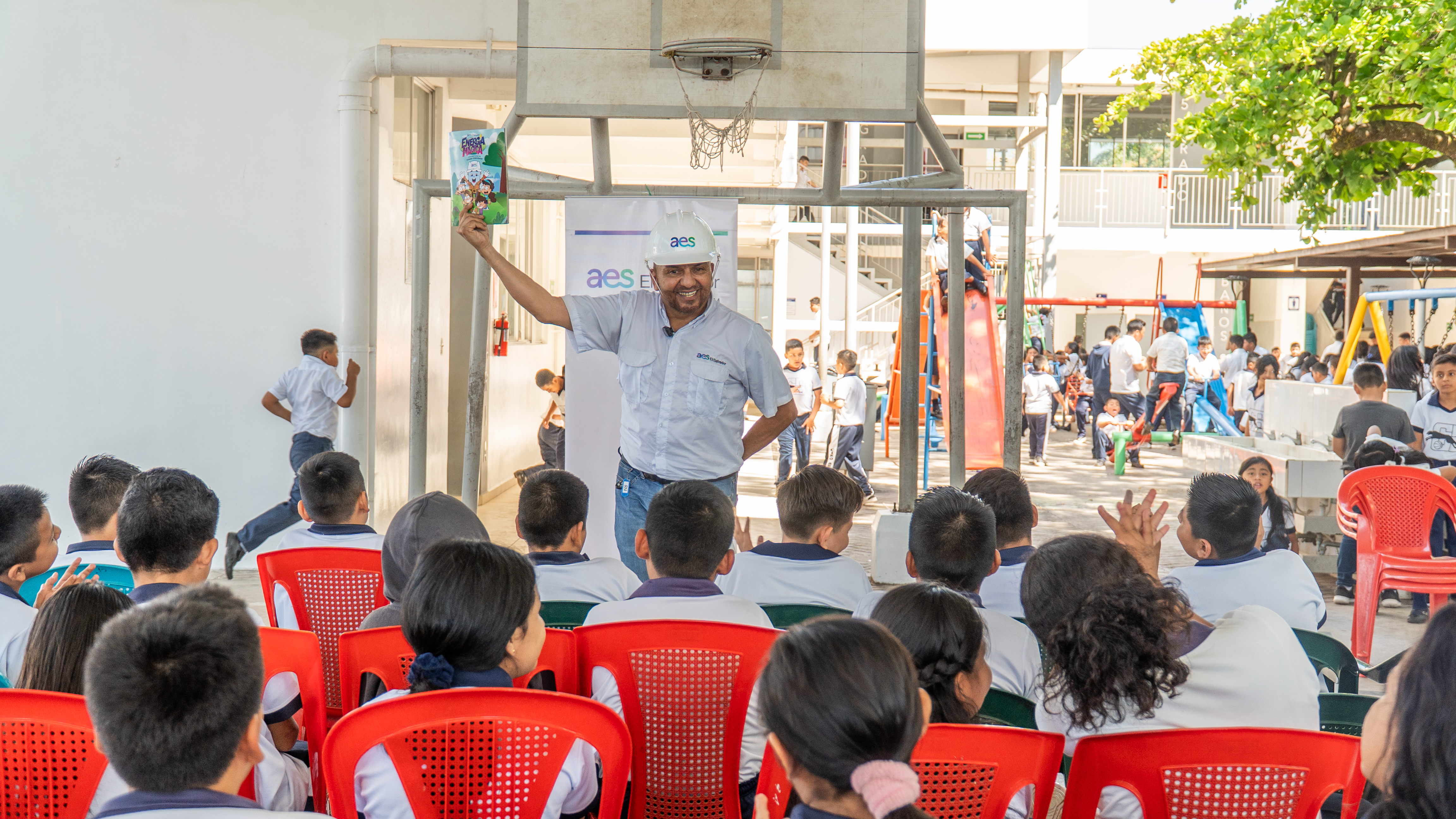 A man wearing a white hard hat and uniform stands in front of a group of seated children in a school courtyard, holding up a colorful booklet. The children are facing him, and other students play on playground equipment in the background.