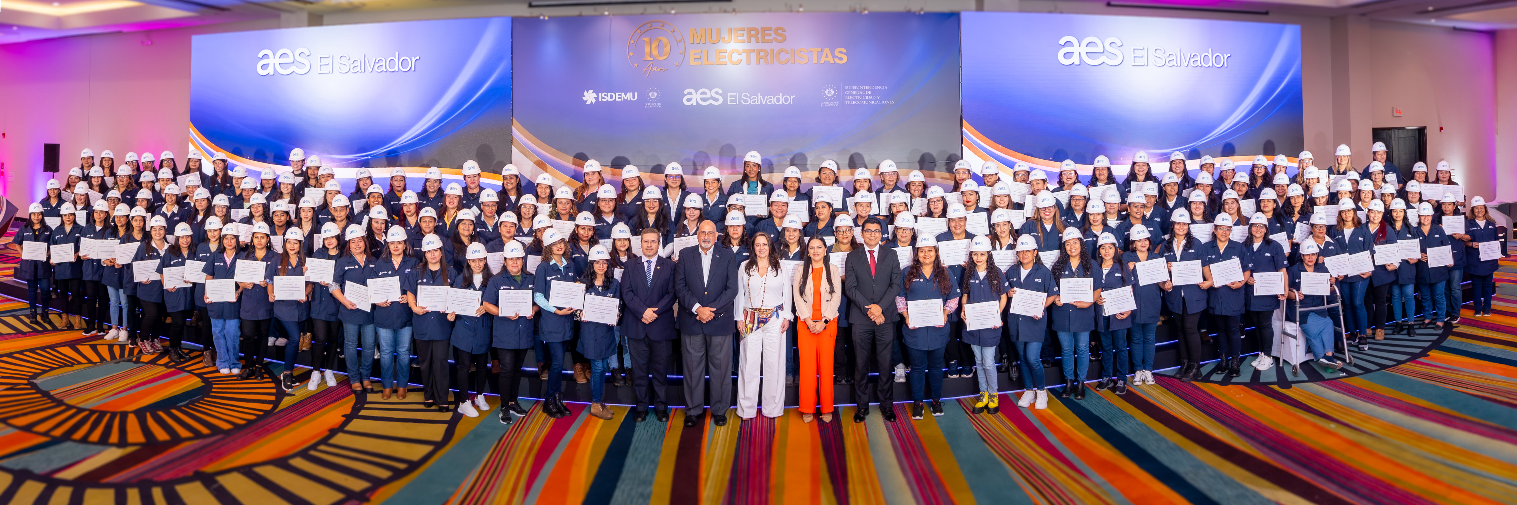 Large group of women in blue uniforms and white helmets holding certificates, standing on a colorful carpeted stage at an AES El Salvador event celebrating 'Women Electricians' 10th anniversary.