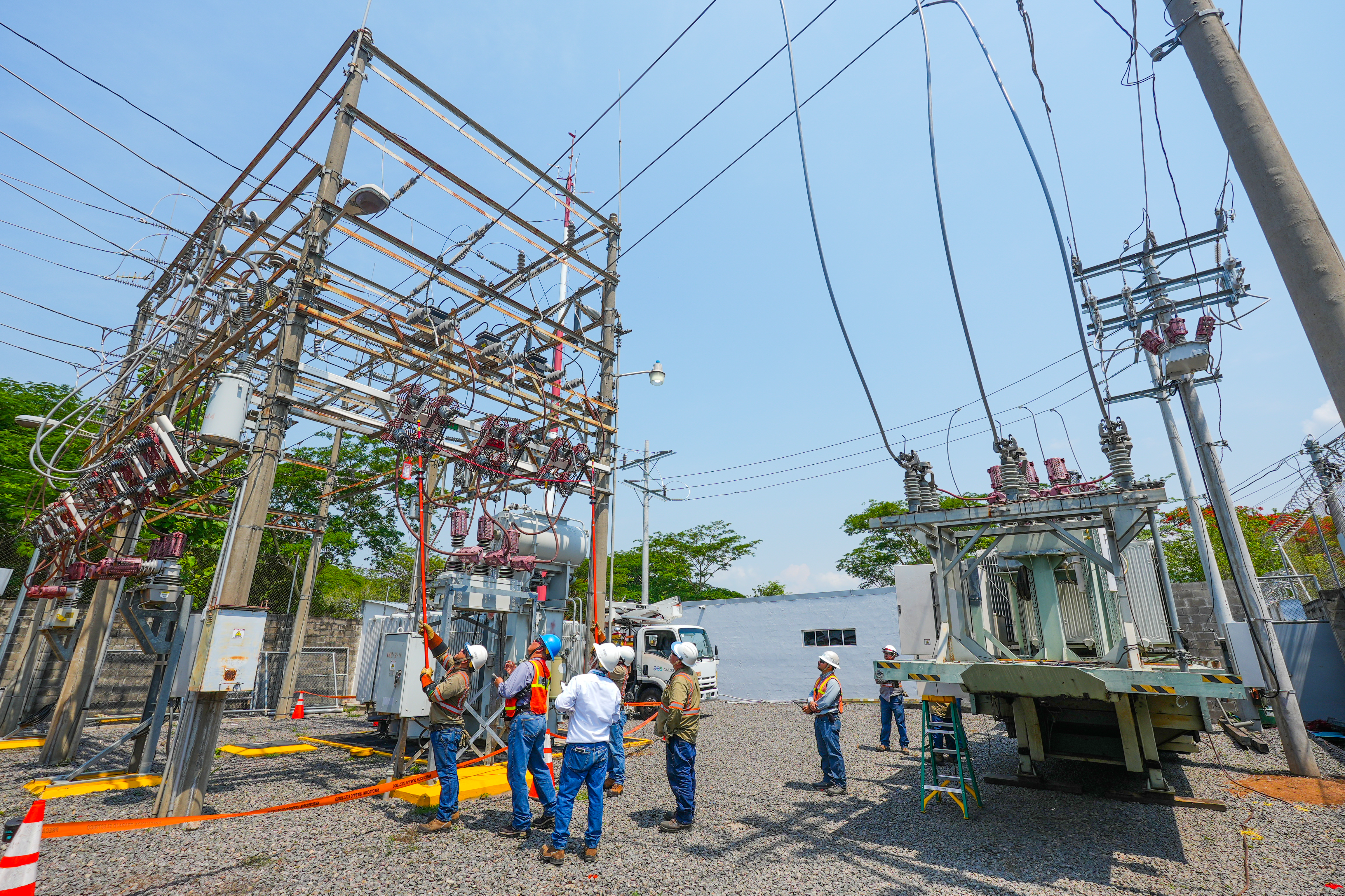 A group of workers wearing safety gear are inspecting electrical equipment at a power substation. The scene includes high-voltage wires, transformers, and utility poles under a clear blue sky.