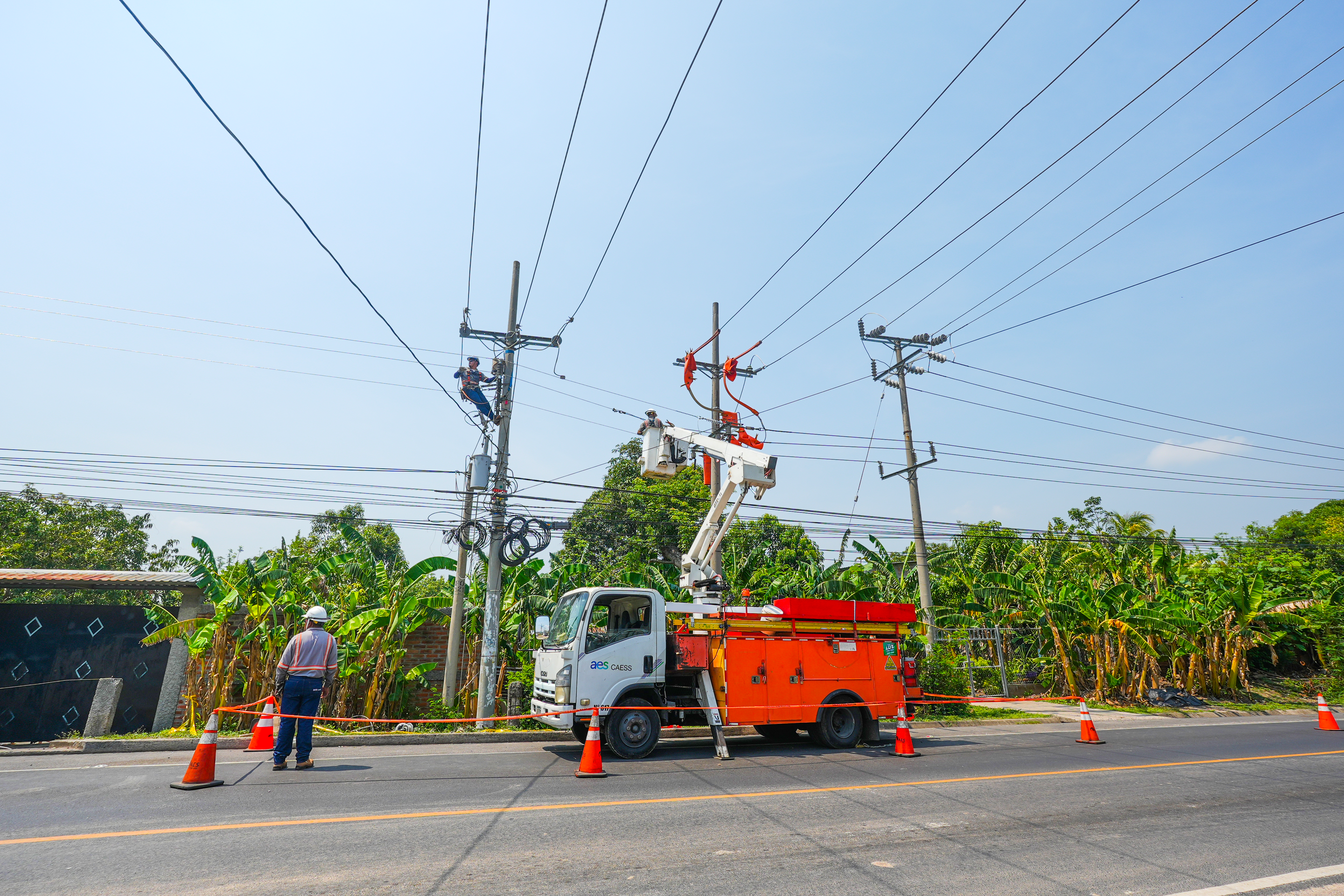 Utility workers repairing power lines on poles with an orange service truck and safety cones on a sunny day.