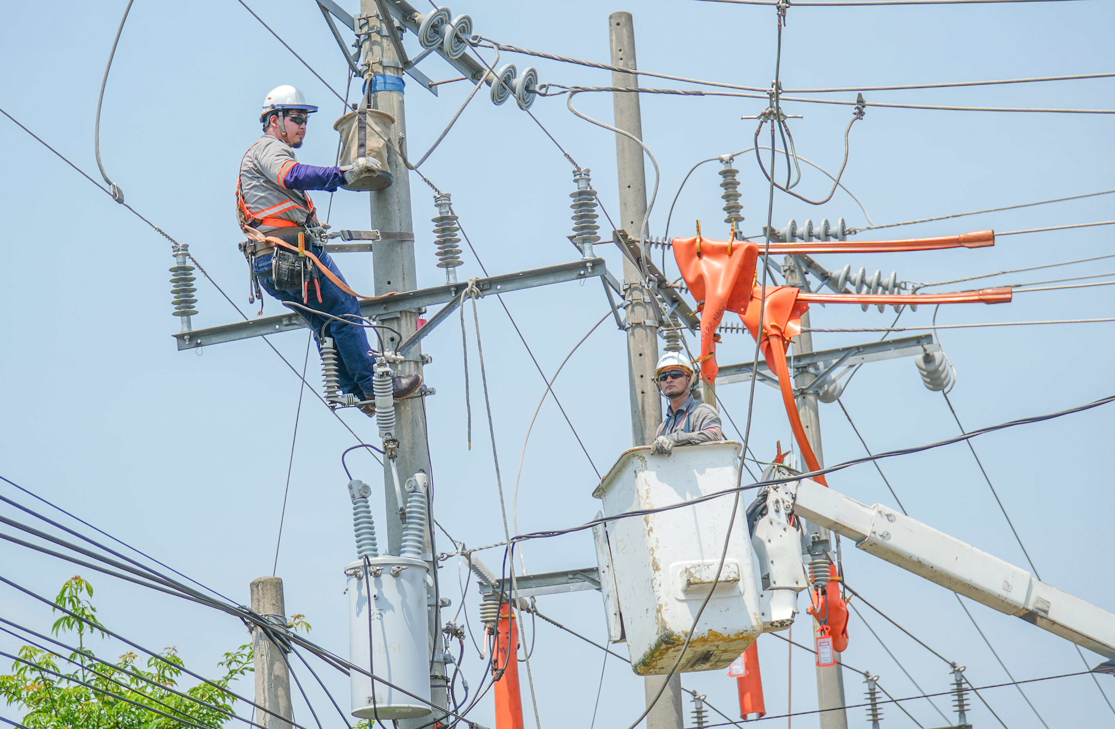 Two utility workers in safety gear are repairing power lines on an electric pole. One worker is climbing the pole while the other is in a bucket lift. The sky is clear and blue.