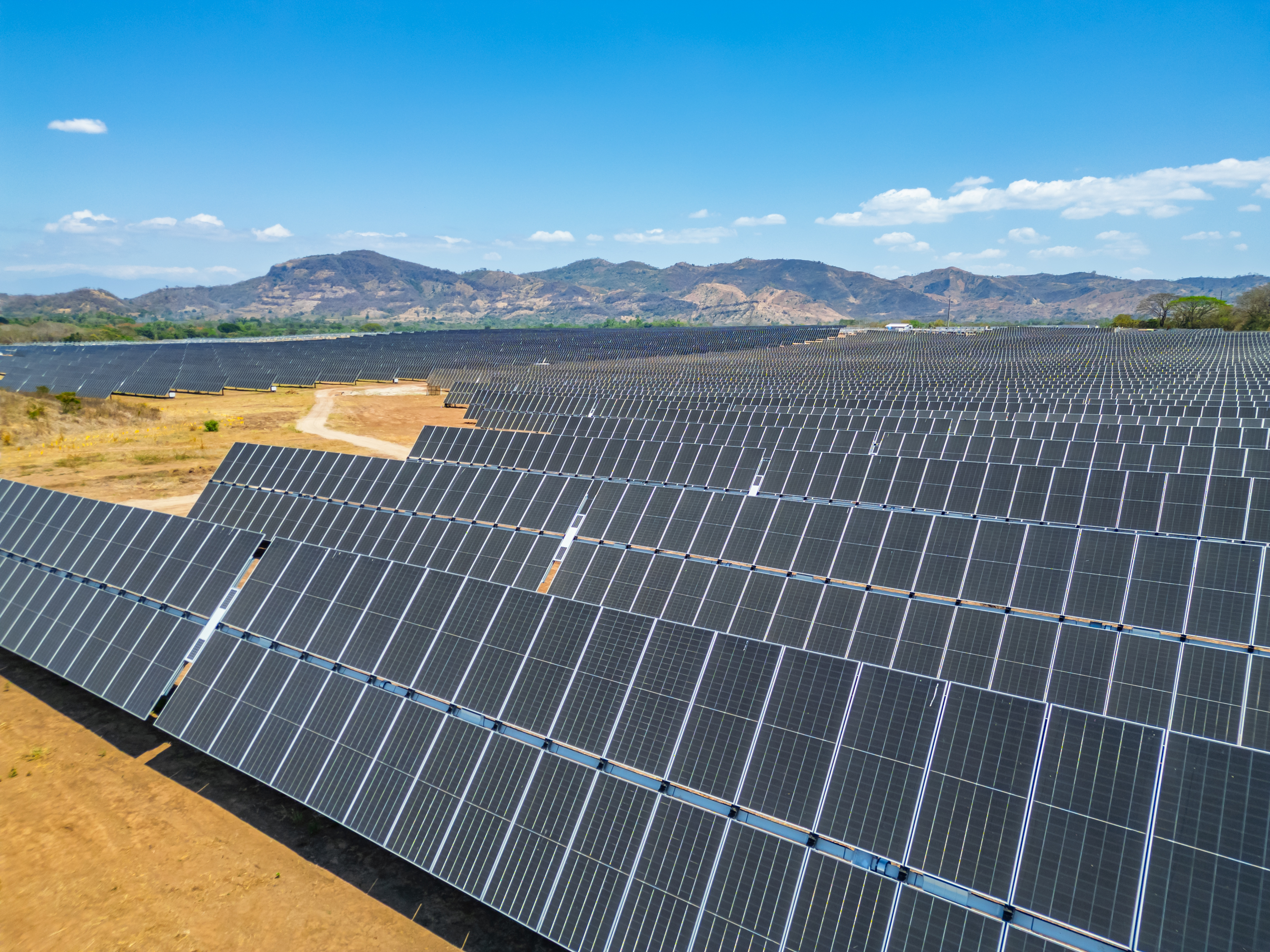 A vast solar farm with rows of solar panels under a clear blue sky, with mountains in the background.