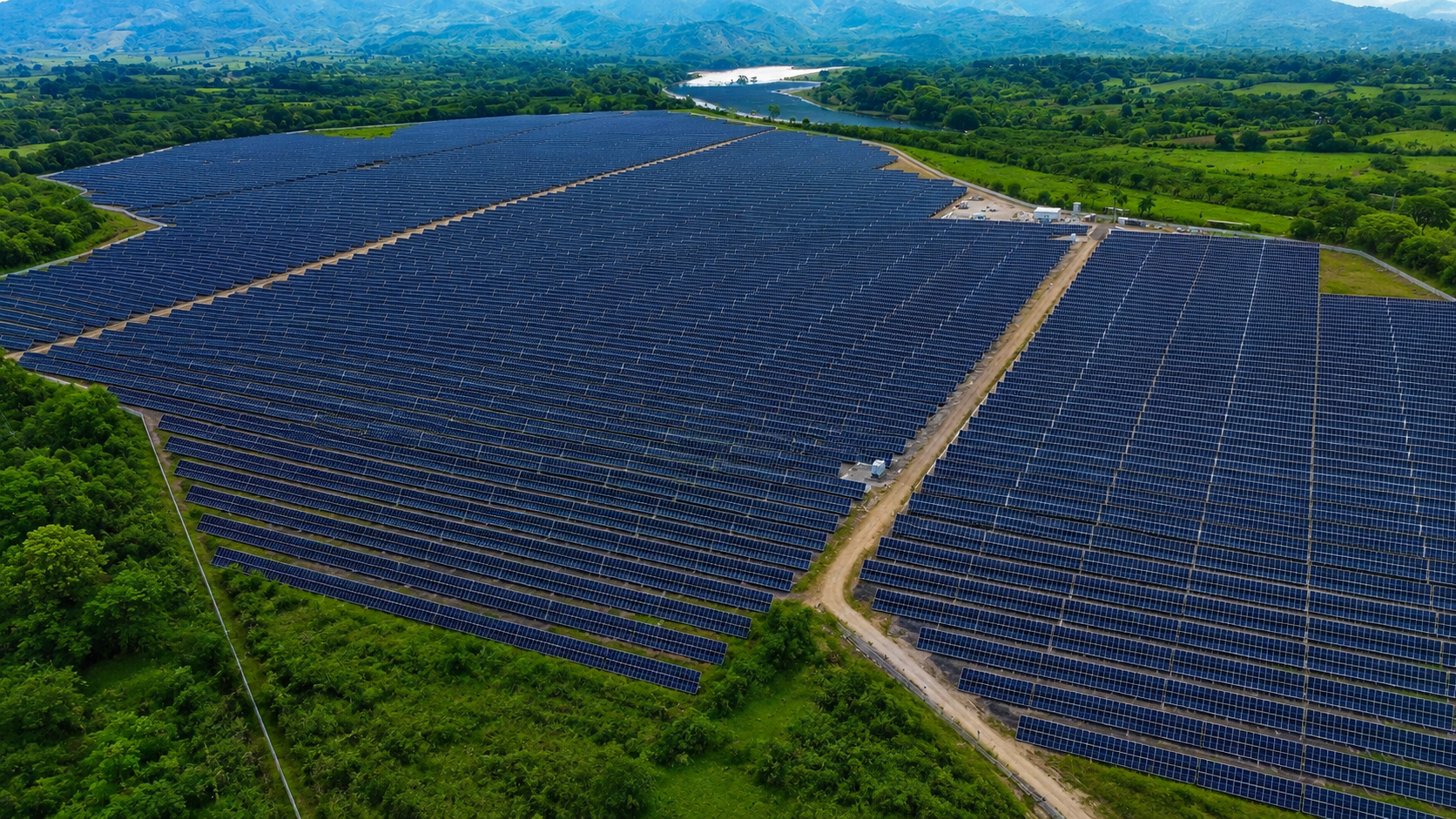 Aerial view of a large solar farm with numerous solar panels arranged in rows, surrounded by lush green vegetation and distant mountains.