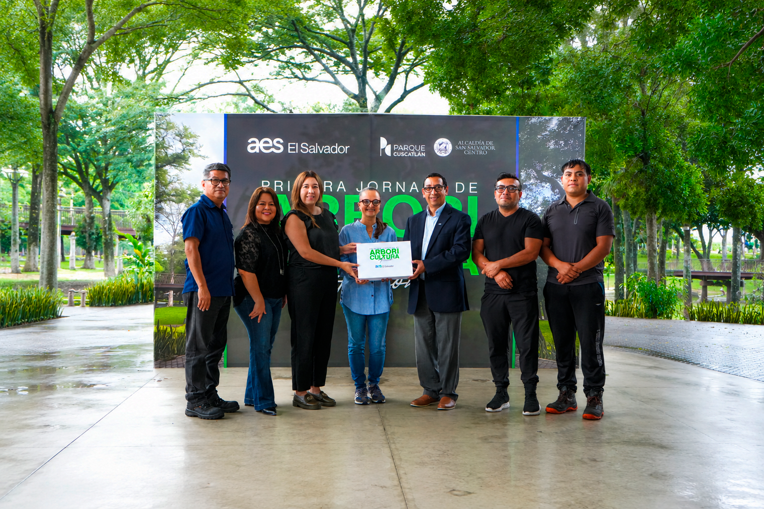 A group of seven people stand in front of a sign at a park event in El Salvador. The sign displays logos and text related to the event. Trees and greenery are visible in the background.
