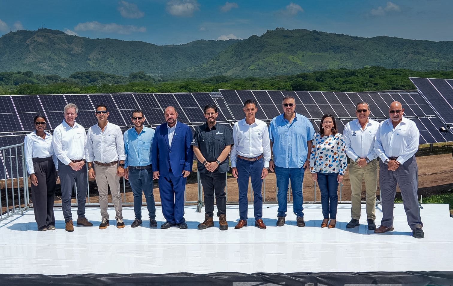 A group of eleven people standing in front of solar panels with a mountainous landscape in the background, all wearing business or casual attire, under a clear blue sky.