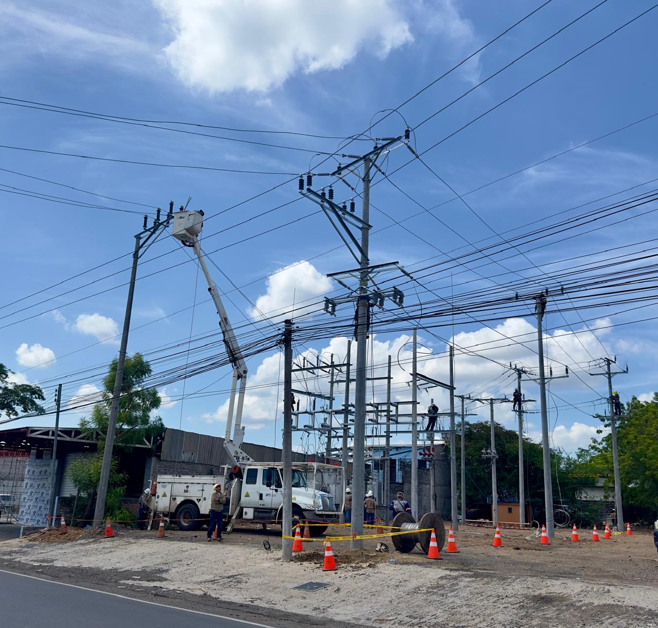 Trabajadores de servicios públicos reparan líneas eléctricas usando un camión con cesta en un día soleado. Varios trabajadores están en el suelo y elevados, rodeados de conos de tráfico y cinta de precaución. El cielo está despejado con nubes dispersas.