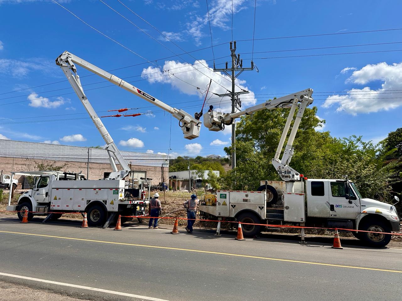 Dos camiones de servicios públicos con elevadores de brazo extendidos están estacionados al lado de la carretera. Los trabajadores usan los elevadores para realizar mantenimiento en las líneas eléctricas. Conos de tráfico y cinta de precaución están colocados alrededor del área por seguridad. El cielo está despejado con algunas nubes.
