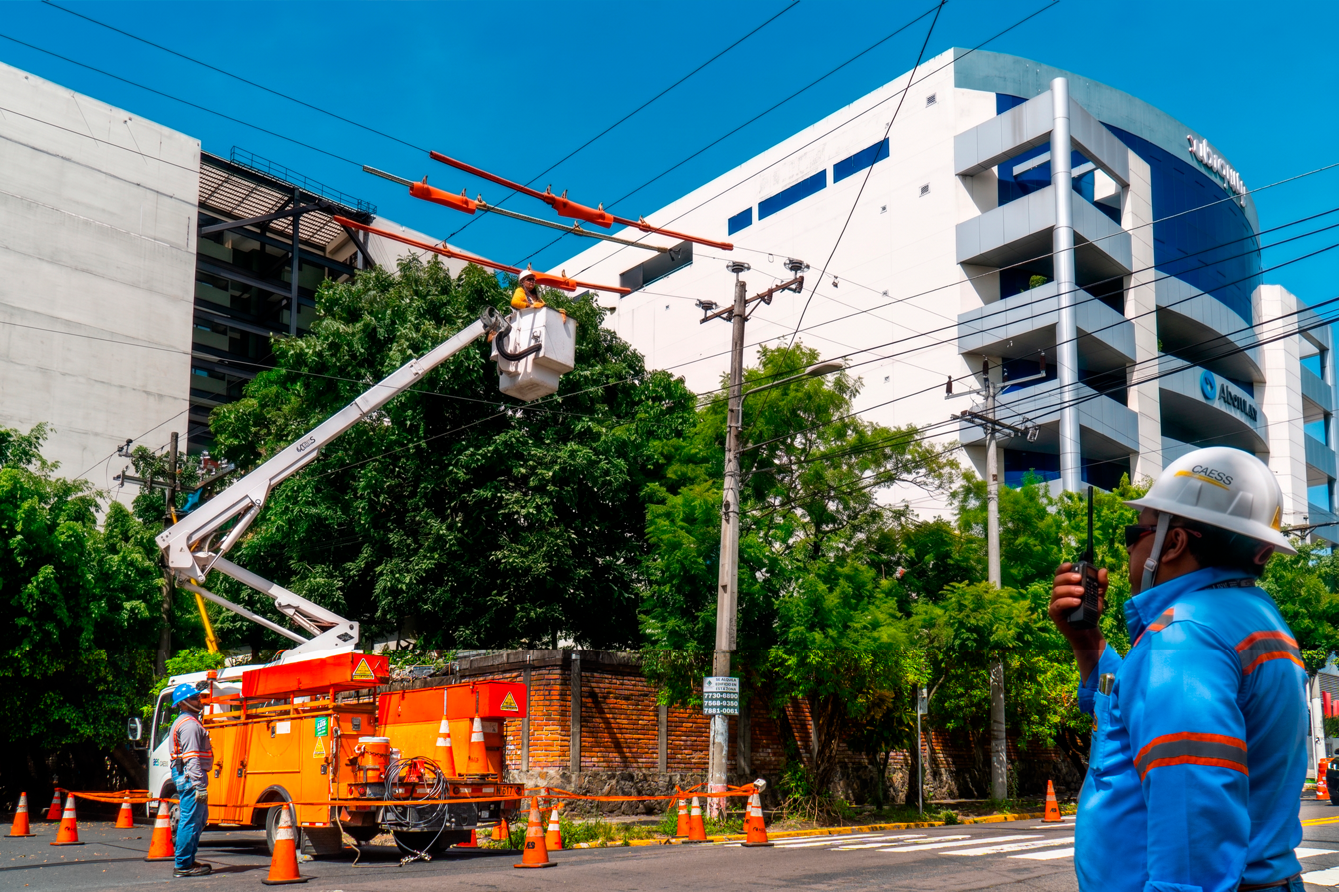 Utility workers repairing power lines using a bucket truck on a city street. One worker is elevated in the bucket, while another communicates via radio. Traffic cones surround the truck for safety.