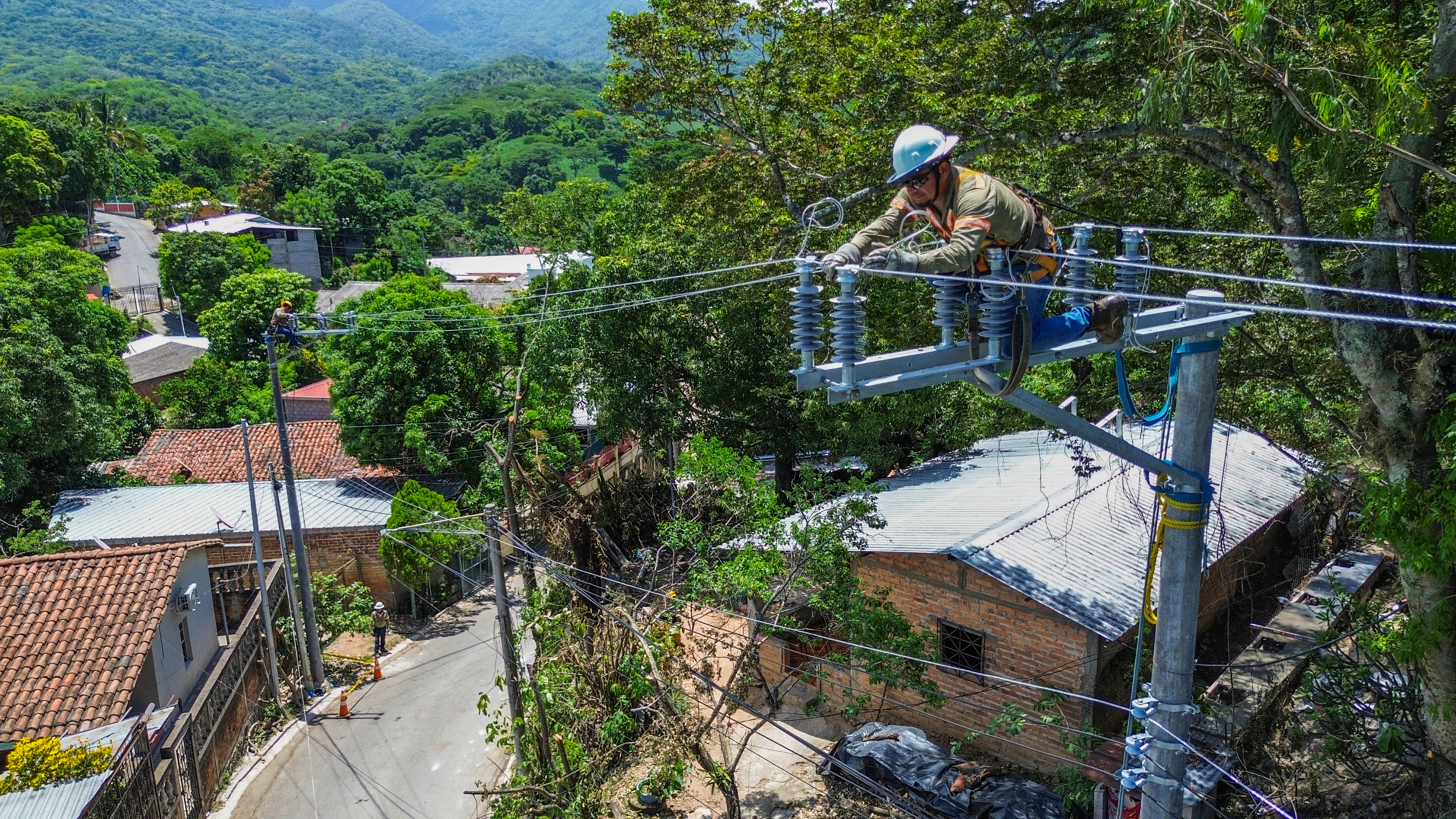 Un-trabajador-con-equipo-de-seguridad-y-casco-blanco-esta-en-lo-alto-de-un-poste-de-concreto - El-hombre-esta-manipulando-un-interruptor-o-equipo-en-las-lineas-electricas-de-alta-tension - En-el-fondo-se-ve-un-pueblo-con-casas-de-techo-de-teja-y-muchos-arboles-verdes-en-una-zona-montañosa - Hay-otro-trabajador-visible-en-el-suelo-cerca-de-la-calle-abajo