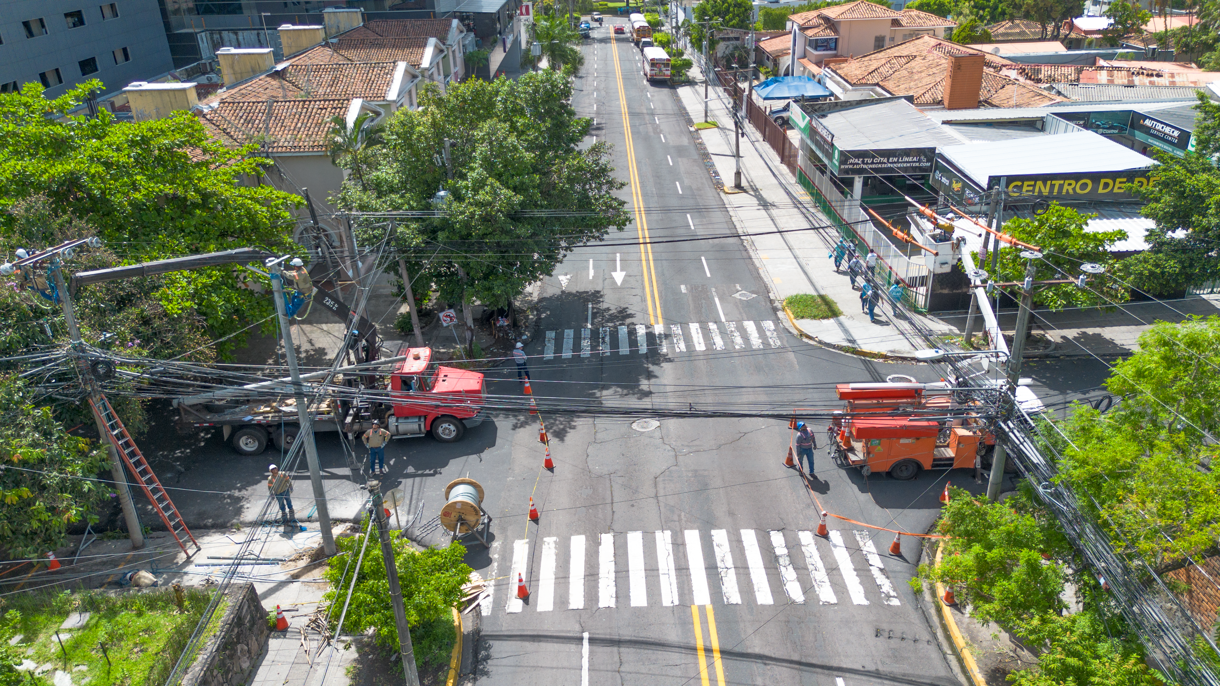 Aerial view of a street with utility workers repairing power lines. Two trucks, one red and one orange, are parked on the road. Workers are on ladders and wearing safety gear. Traffic cones are placed around the work area, and trees line the street.