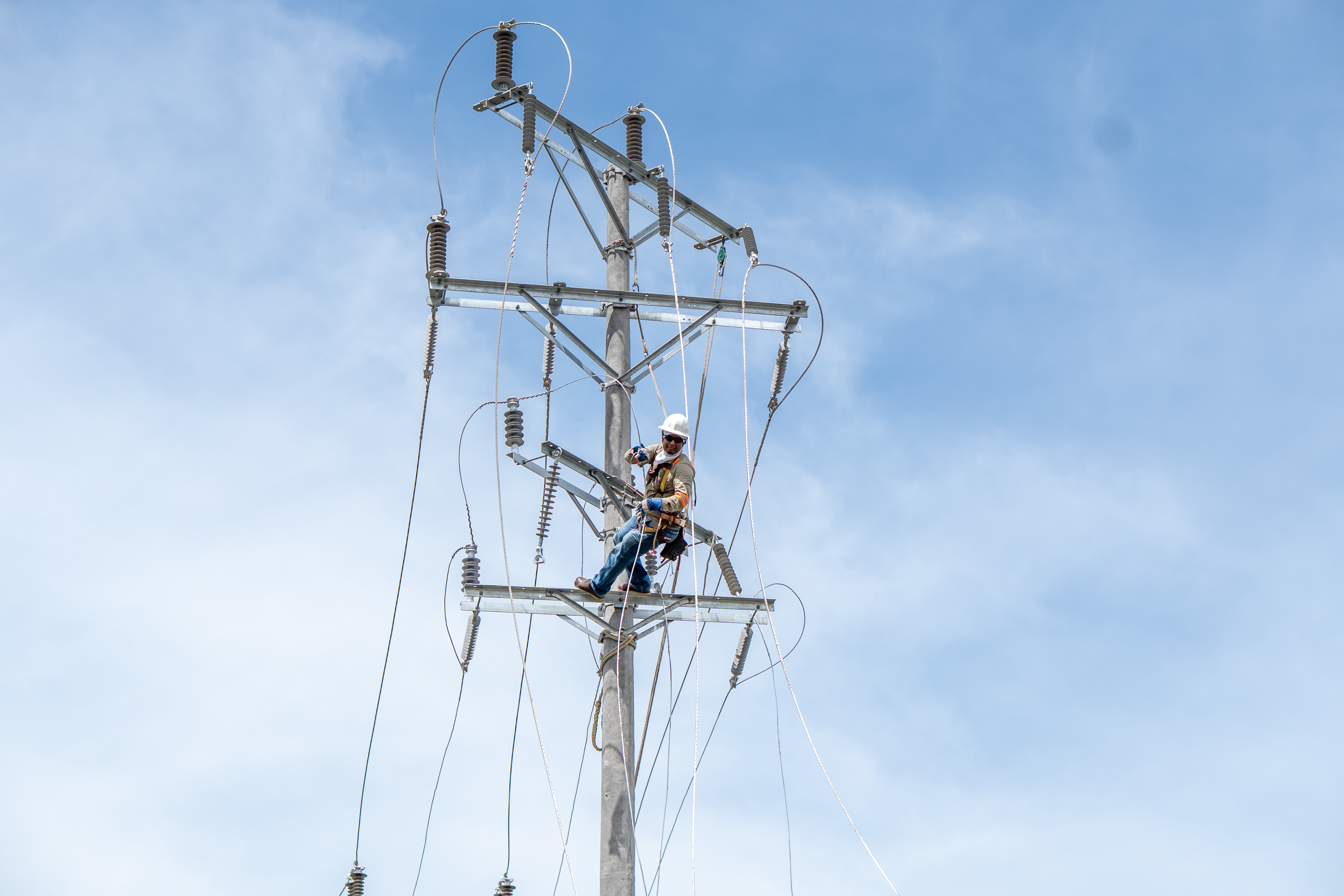 Un técnico liniero equipado con casco blanco, arnés de seguridad y gafas oscuras realiza labores de mantenimiento o instalación en lo alto de un poste de luz de alta tensión. El poste de hormigón cuenta con una estructura metálica llena de aislantes de porcelana, cables eléctricos y cuerdas de sujeción. Al fondo se observa un cielo despejado con ligeras nubes.
