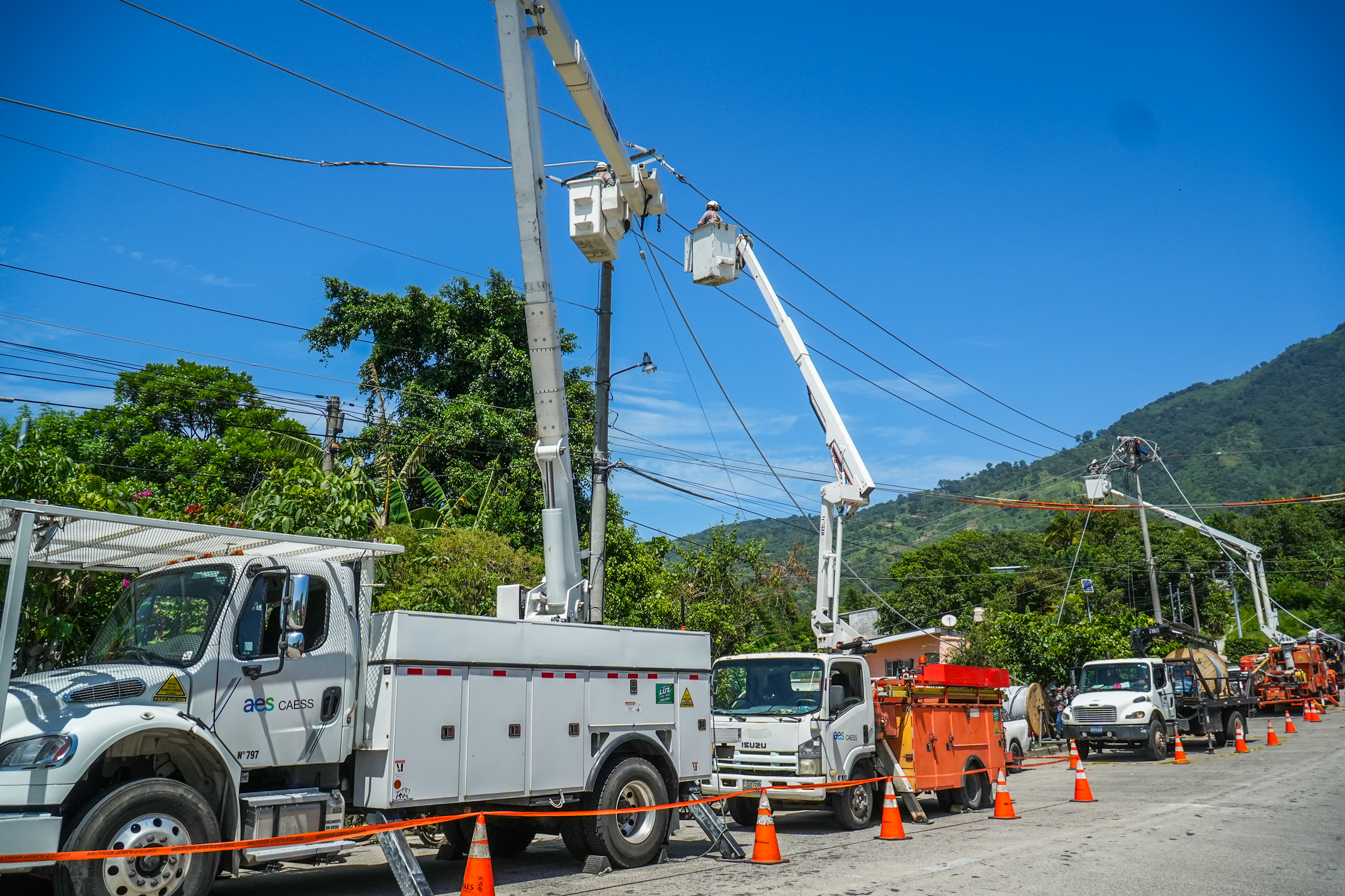Varios operarios trabajan en altura dentro de las canastas elevadas de dos camiones grúa, manipulando los cables de tensión. La zona está delimitada por conos de tráfico naranjas. De fondo hay una montaña cubierta de vegetación bajo un cielo azul despejado.