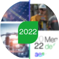 A classroom with students seated at wooden desks facing a whiteboard. The teacher stands at the front, pointing to the board with text and diagrams. Another adult sits at a corner desk. The walls feature educational posters and a "Bienvenidos" sign above the whiteboard.