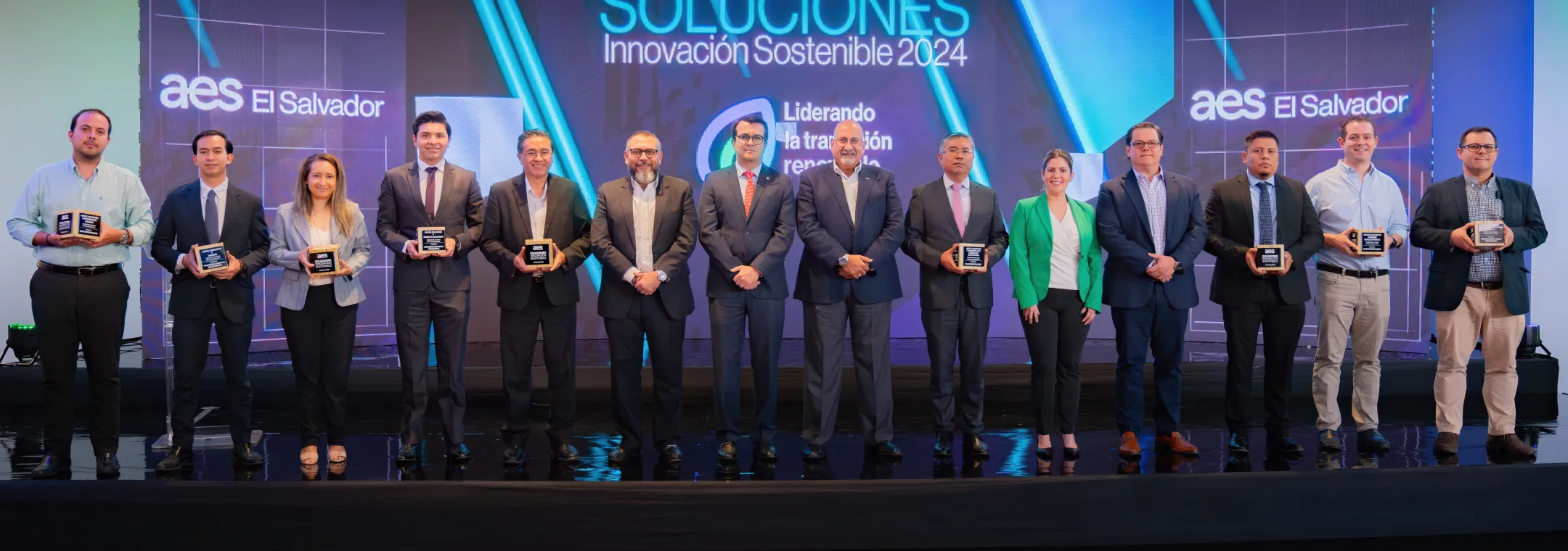 A group of 14 individuals standing on stage at the AES El Salvador Sustainable Solutions 2024 event. Each person is holding an award plaque, symbolizing recognition for their contributions. The background features a vibrant event banner with the words "Soluciones Innovación Sostenible 2024." The group is dressed in business attire, highlighting a formal and celebratory occasion.