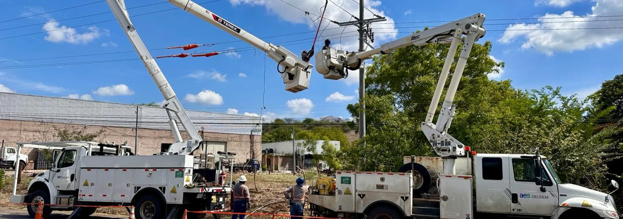 Dos camiones de servicios públicos con elevadores de brazo extendidos están estacionados al lado de la carretera. Los trabajadores usan los elevadores para realizar mantenimiento en las líneas eléctricas. Conos de tráfico y cinta de precaución están colocados alrededor del área por seguridad. El cielo está despejado con algunas nubes.
