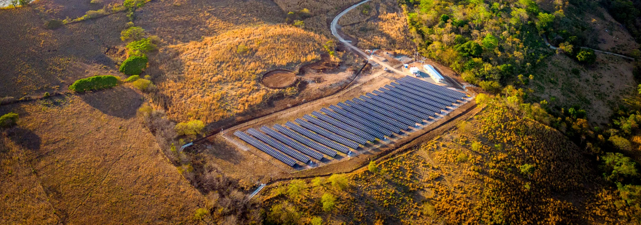 Aerial view of a solar farm situated on a hilly landscape near a large body of water. The hills are covered with patches of green vegetation and dry grass, with a winding road leading to the solar panels. In the background, a calm blue sea stretches towards distant mountains under a clear sky.