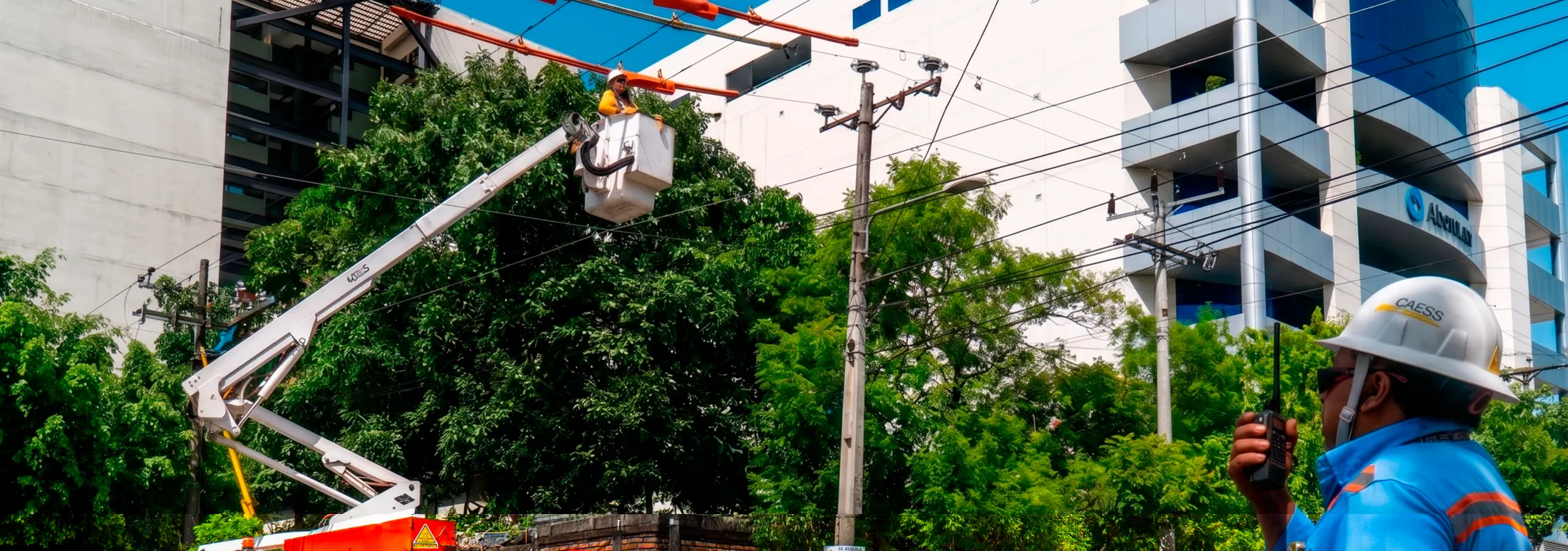 Utility workers repairing power lines using a bucket truck on a city street. One worker is elevated in the bucket, while another communicates via radio. Traffic cones surround the truck for safety.