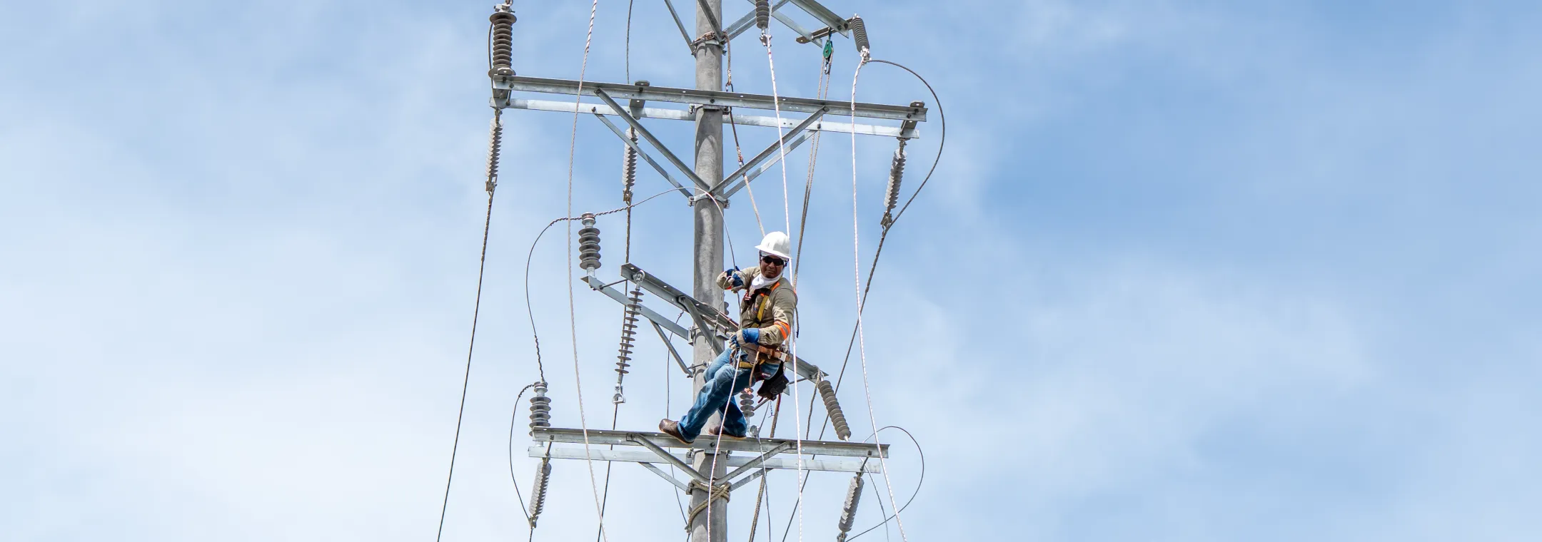 Un técnico liniero equipado con casco blanco, arnés de seguridad y gafas oscuras realiza labores de mantenimiento o instalación en lo alto de un poste de luz de alta tensión. El poste de hormigón cuenta con una estructura metálica llena de aislantes de porcelana, cables eléctricos y cuerdas de sujeción. Al fondo se observa un cielo despejado con ligeras nubes.