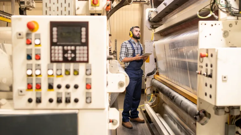A factory worker wearing blue overalls and a plaid shirt stands in front of a large industrial machine, inspecting the equipment while holding a tablet. He wears protective yellow earmuffs. In the foreground, a control panel with multiple buttons and switches is visible. The scene highlights the worker's focus on monitoring and operating the machinery in the industrial setting.