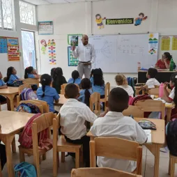 A classroom with students seated at wooden desks facing a whiteboard. The teacher stands at the front, pointing to the board with text and diagrams. Another adult sits at a corner desk. The walls feature educational posters and a "Bienvenidos" sign above the whiteboard.