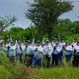 A group of people wearing white shirts and green caps stand outdoors surrounded by trees and greenery. Many have one arm raised, suggesting a group gesture or celebration.